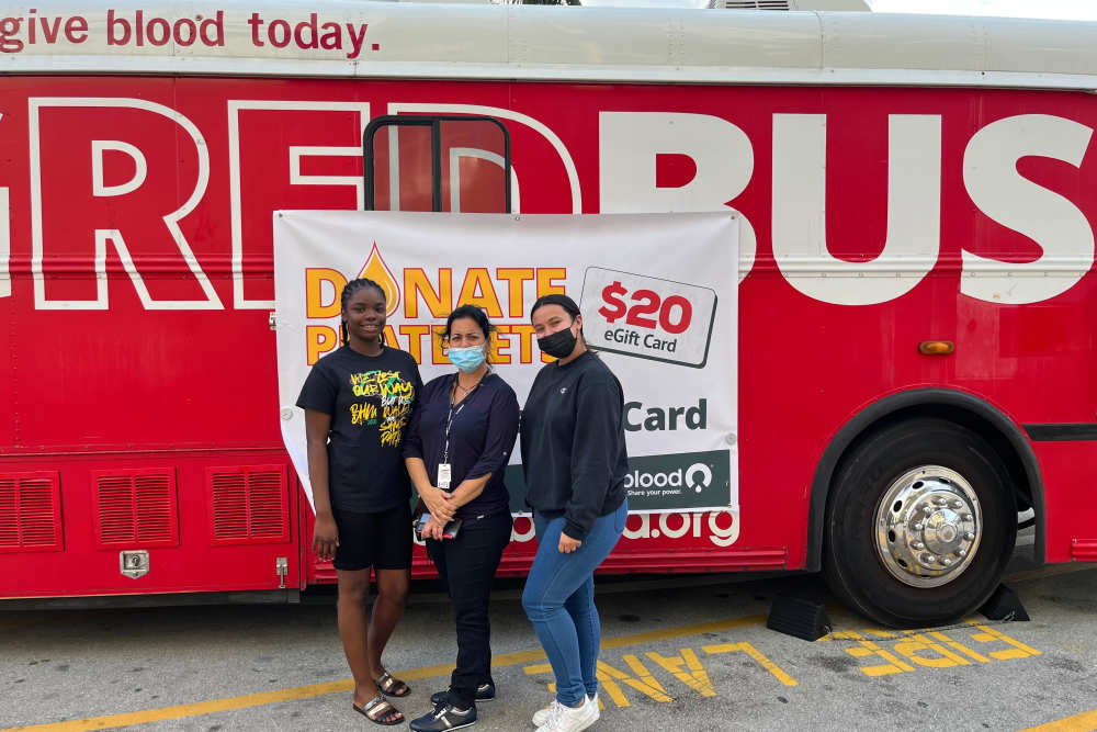 Three women standing in front of a red blood donation bus with a donation banner at Bayview Student Living in North Miami, Florida