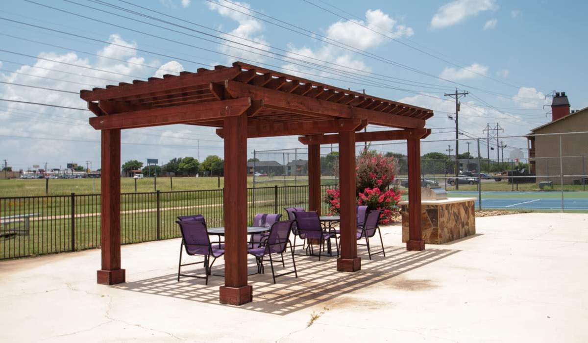 Wooden pergola with seating area and outdoor grill surrounded by green grass and fencing at Riatta Ranch Apartments in Abilene, Texas