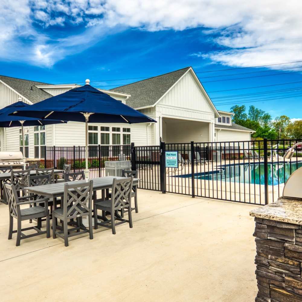 Outdoor patio area with dining sets and umbrellas near a swimming pool and grill, against a backdrop of a white building and blue sky at Commonwealth Apartments in Midlothian, Virginia
