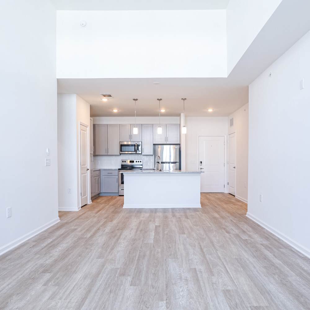 A modern kitchen and living area with light wood flooring, white walls, stainless steel appliances, and a central island with pendant lights above at Commonwealth Apartments in Midlothian, Virginia
