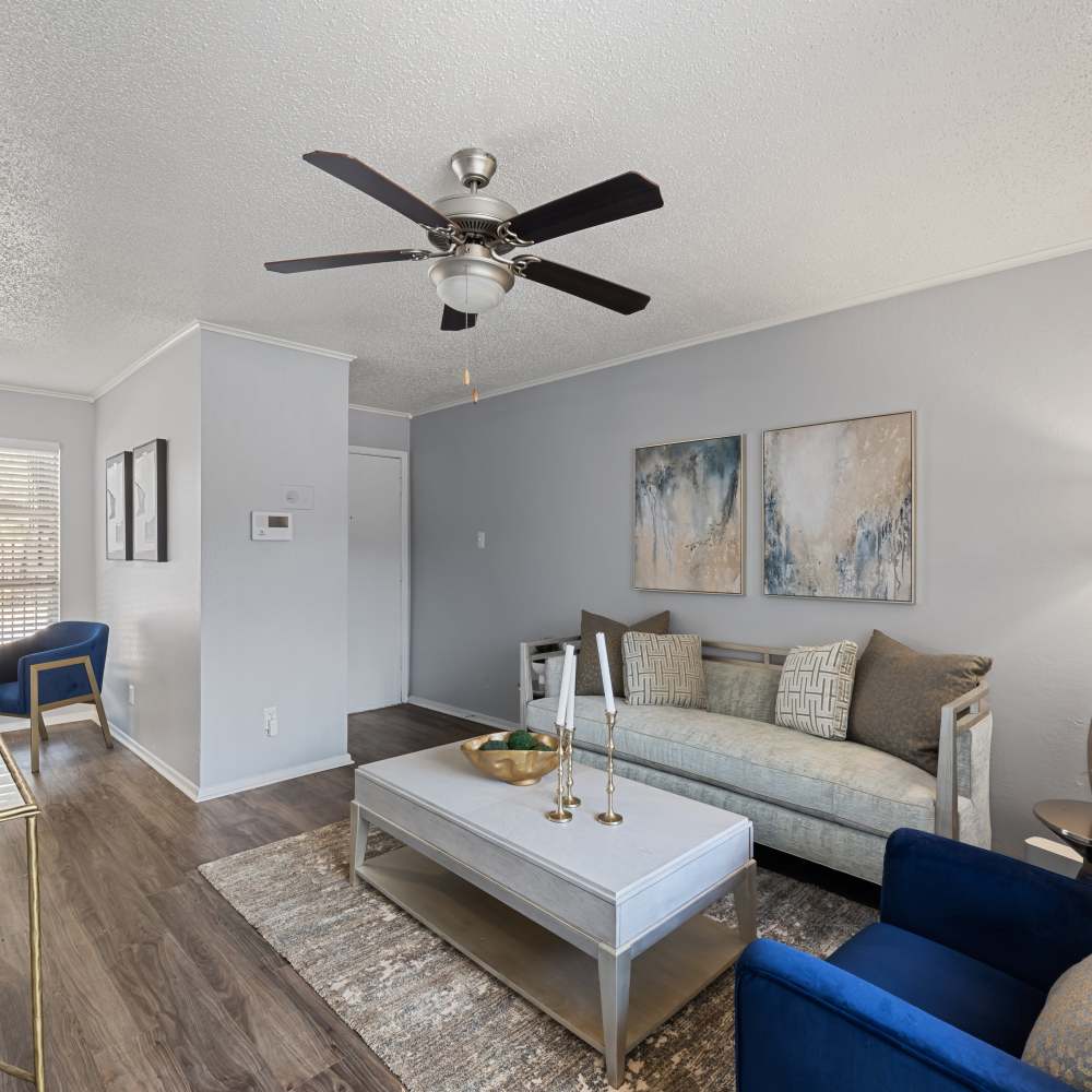 Inviting living room with a light gray sofa, blue accent chairs, a coffee table, and abstract artwork, featuring a ceiling fan at The Garrison in Dallas, Texas