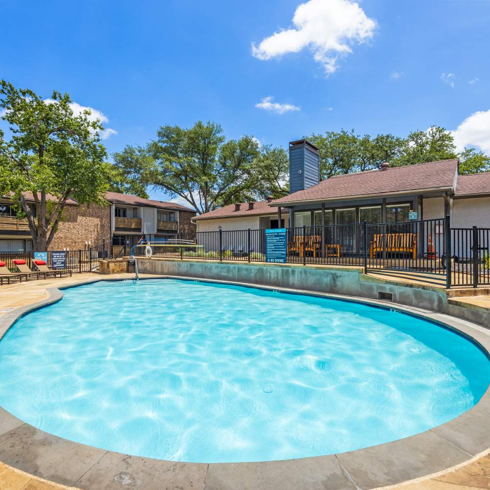View of a curved swimming pool with clear water, surrounded by lounge chairs and apartment buildings, under a bright blue sky at The Garrison in Dallas, Texas