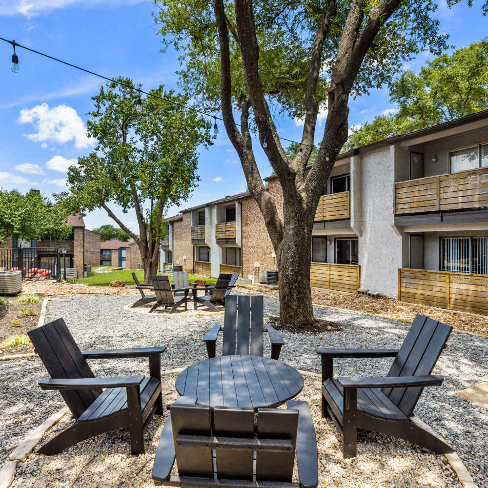 Outdoor communal area with seating around a fire pit, featuring gravel landscaping and surrounded by apartment buildings under a sunny blue sky at The Garrison in Dallas, Texas
