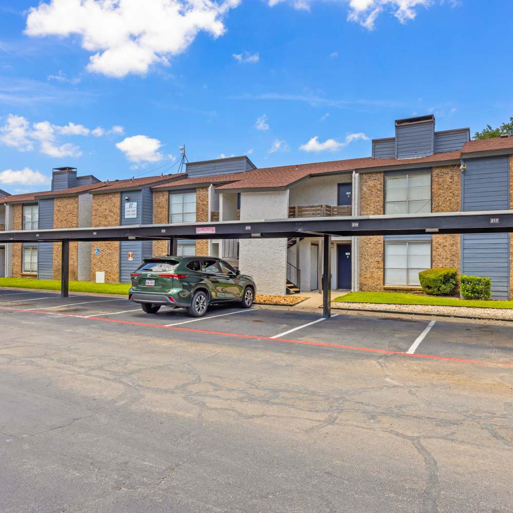 Residential apartment complex with brick and siding exterior, covered parking spaces, single car, and trees in background at The Garrison in Dallas, Texas
