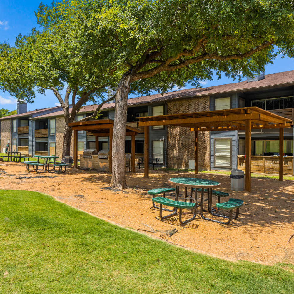 Outdoor seating area with picnic tables and benches under a wooden pergola, surrounded by grass and trees, in front of a two-story apartment building at The Garrison in Dallas, Texas