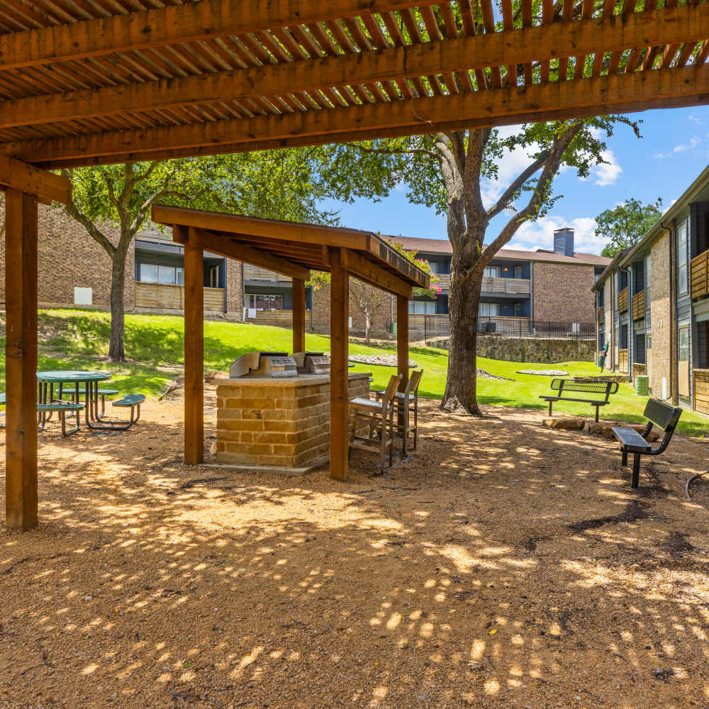 Outdoor area with wooden pergola over a barbecue grill station, surrounded by chairs and tables, set in a grassy courtyard near apartment buildings, under a clear blue sky at The Garrison in Dallas, Texas