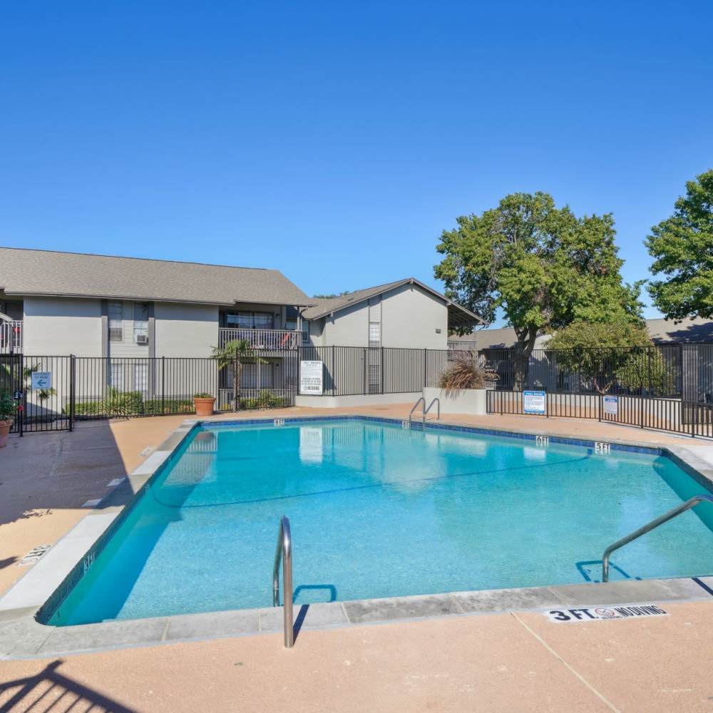 Sparkling pool surrounded by lush greenery and sunlit clubhouse at Woodlands of Plano in Plano, Texas
