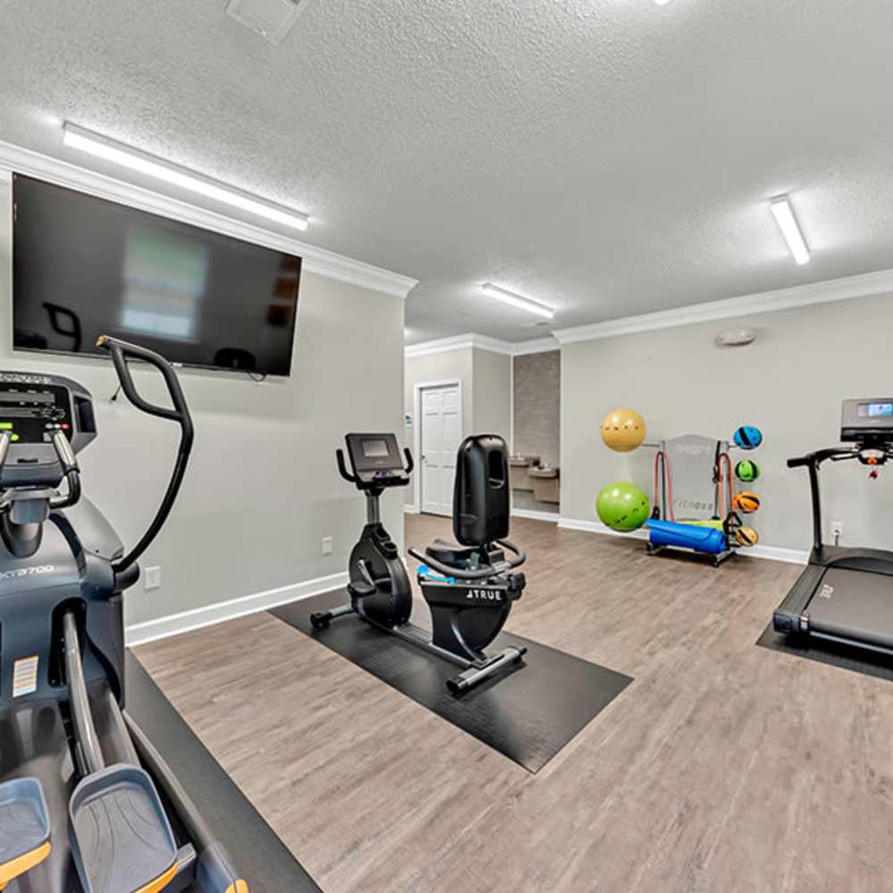 Modern fitness room with elliptical trainer, recumbent bike, treadmill, wall-mounted TV, exercise balls, and yoga mat rack at Osprey Place Apartments in North Charleston, South Carolina  