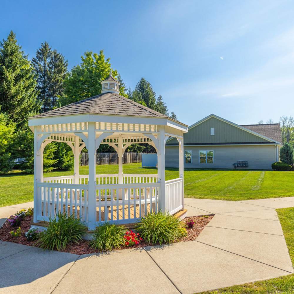 White gazebo surrounded by green grass and colorful flowers under a clear blue sky at Carriages At Cedar Rock in Webster, New York