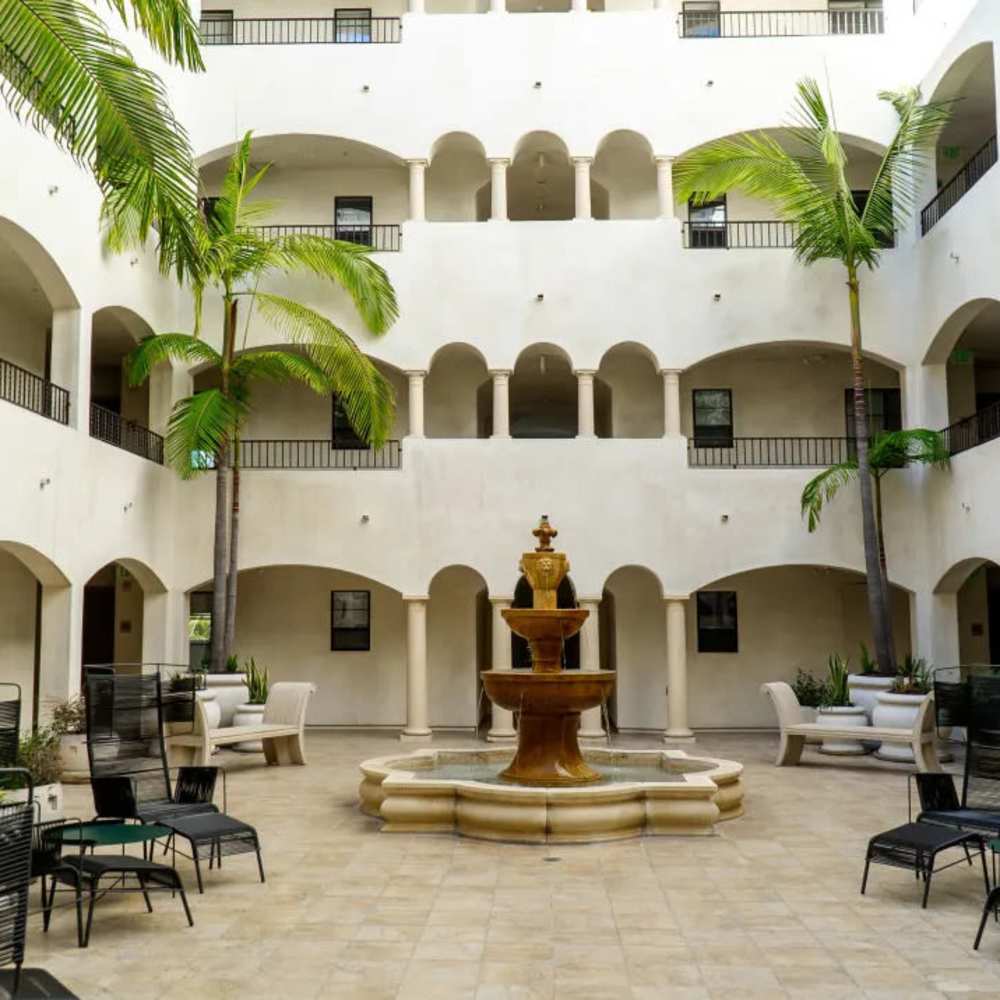 Two-Bedroom Luxury Apartments in Los Angeles, CA - Luxe Villas - Courtyard with a Stone Fountain, Benches, Woven Chairs, Potted Palm Trees, and View of the Building Railings and Arches at Vida Las Vegas in Las Vegas, Nevada