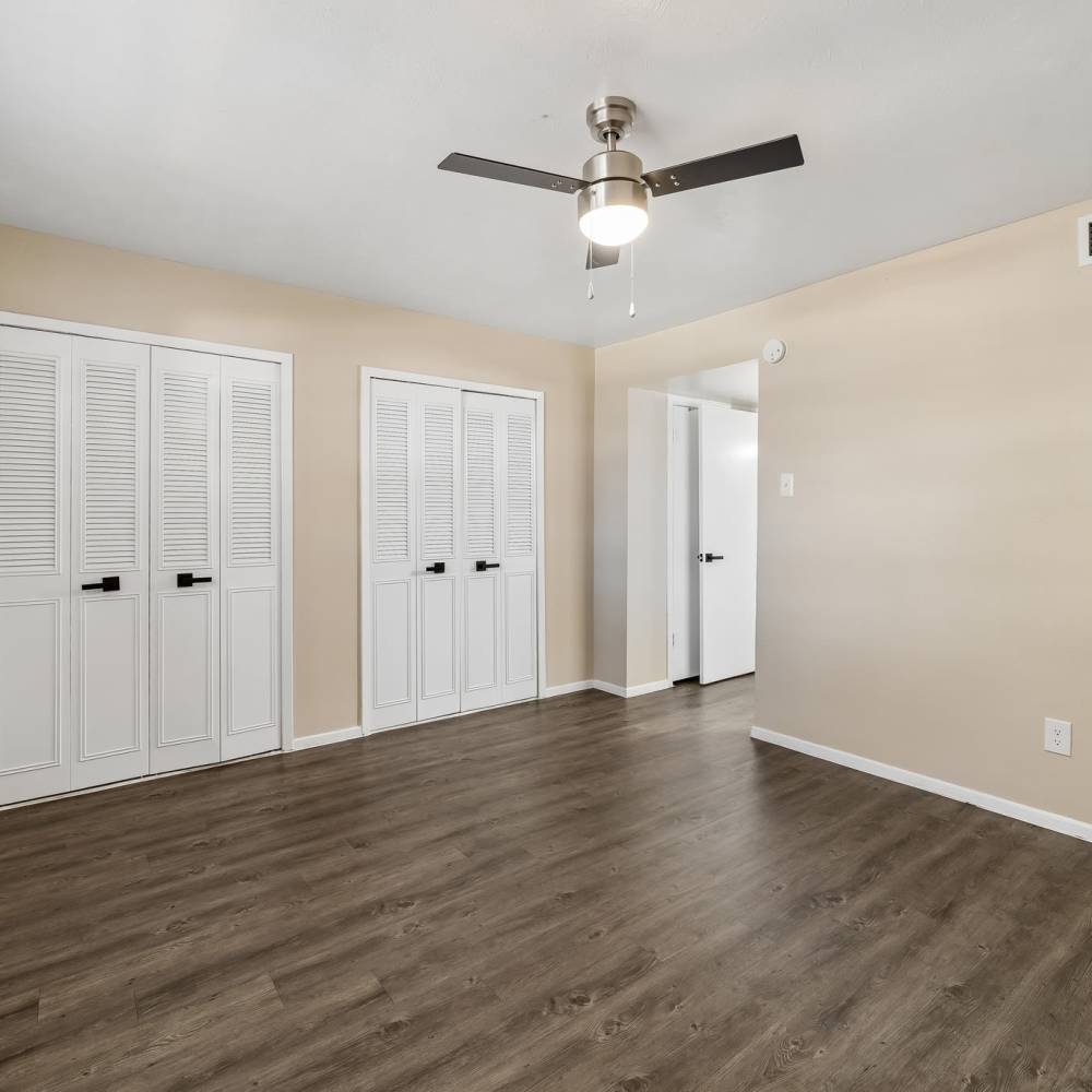 Empty beige bedroom with dark wood flooring, multiple white louvered closets, ceiling fan and light, and an open white door. Clean, bright interior at Royal Arms in Richardson,Texas