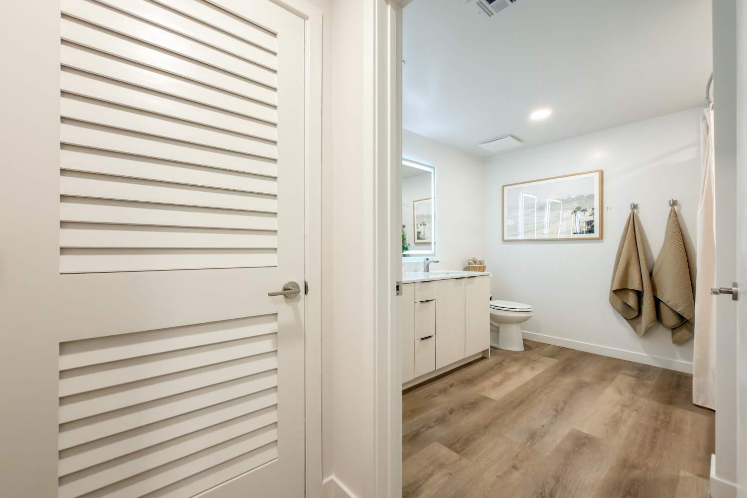 View into bathroom with white vanity, toilet, towels on hooks, and light wood flooring at RIVA Solana Beach in Solana Beach,California