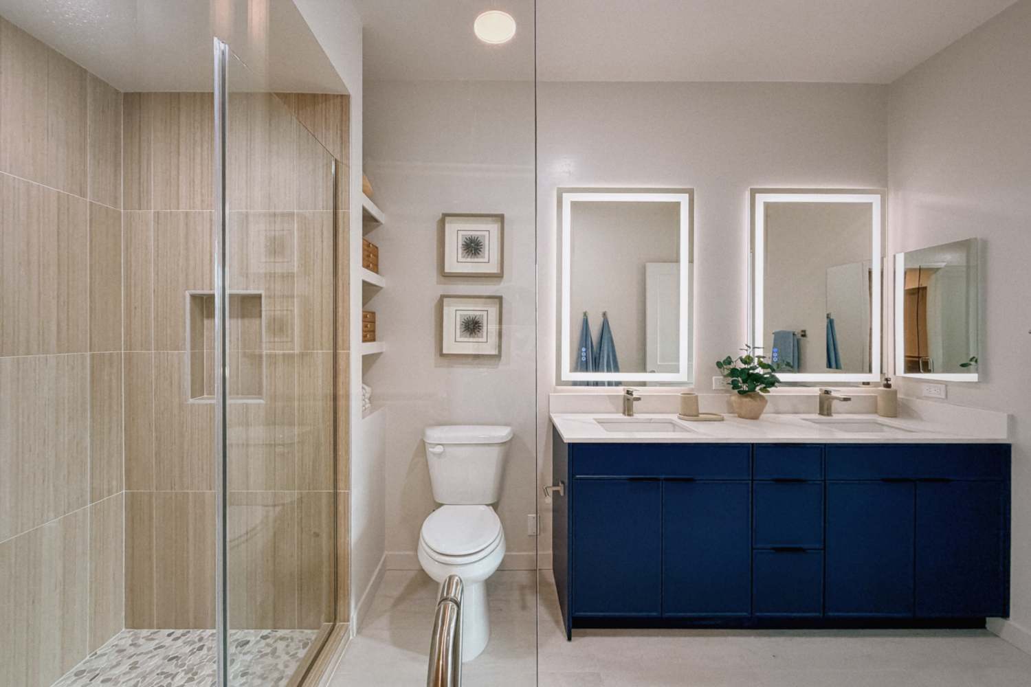 Bathroom with glass‑enclosed shower, toilet, double vanity with blue cabinets, and illuminated mirrors at RIVA Solana Beach in Solana Beach,California