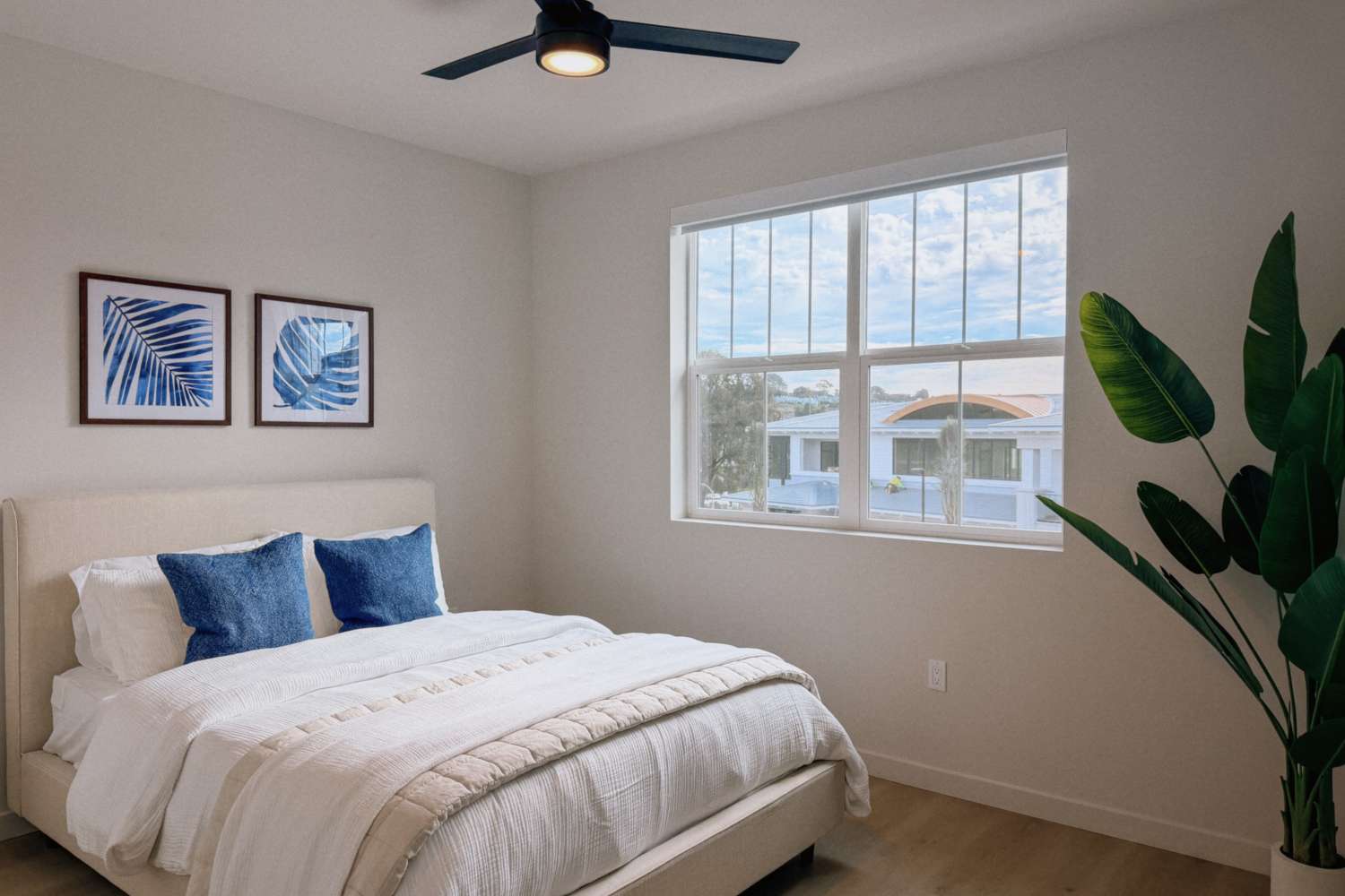 Bright bedroom with a neatly made bed, blue accent pillows, framed wall art, ceiling fan, and large window at RIVA Solana Beach in Solana Beach,California
