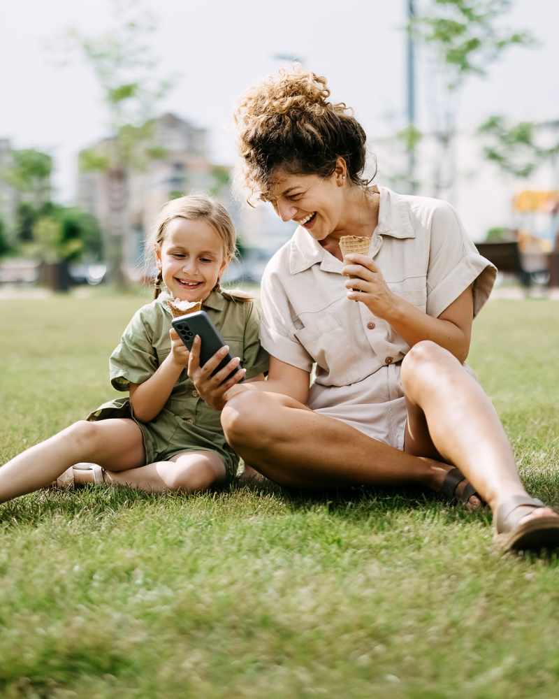 Resident using mobile while enjoying a ice cream with her daughter sitting on green lawn at The Indie Glendale Collection in Glendale, California