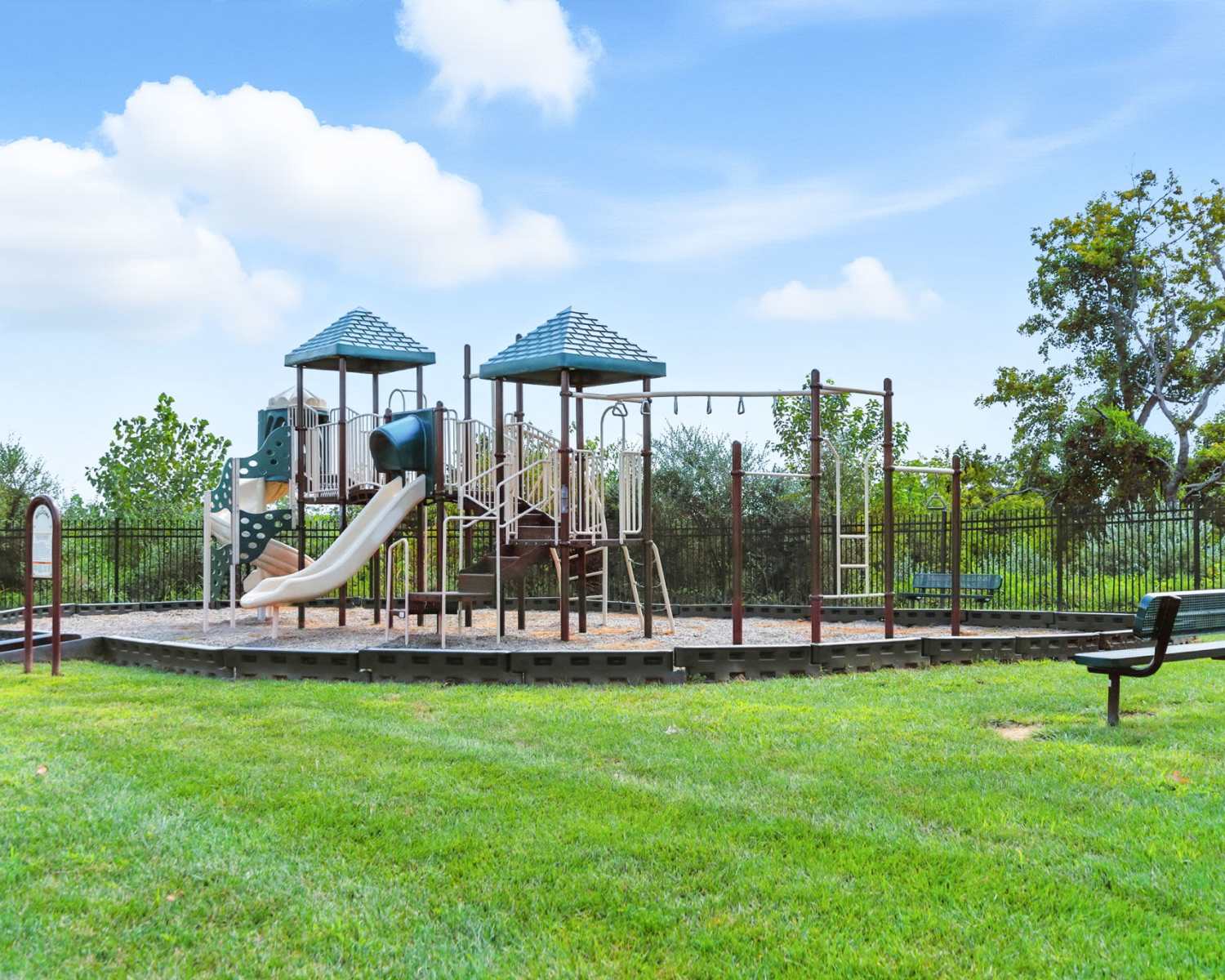 Playground with slides and swings surrounded by green grass and trees, fenced area for safety at Bristol Bluffs Apartments in Louisville, Kentucky