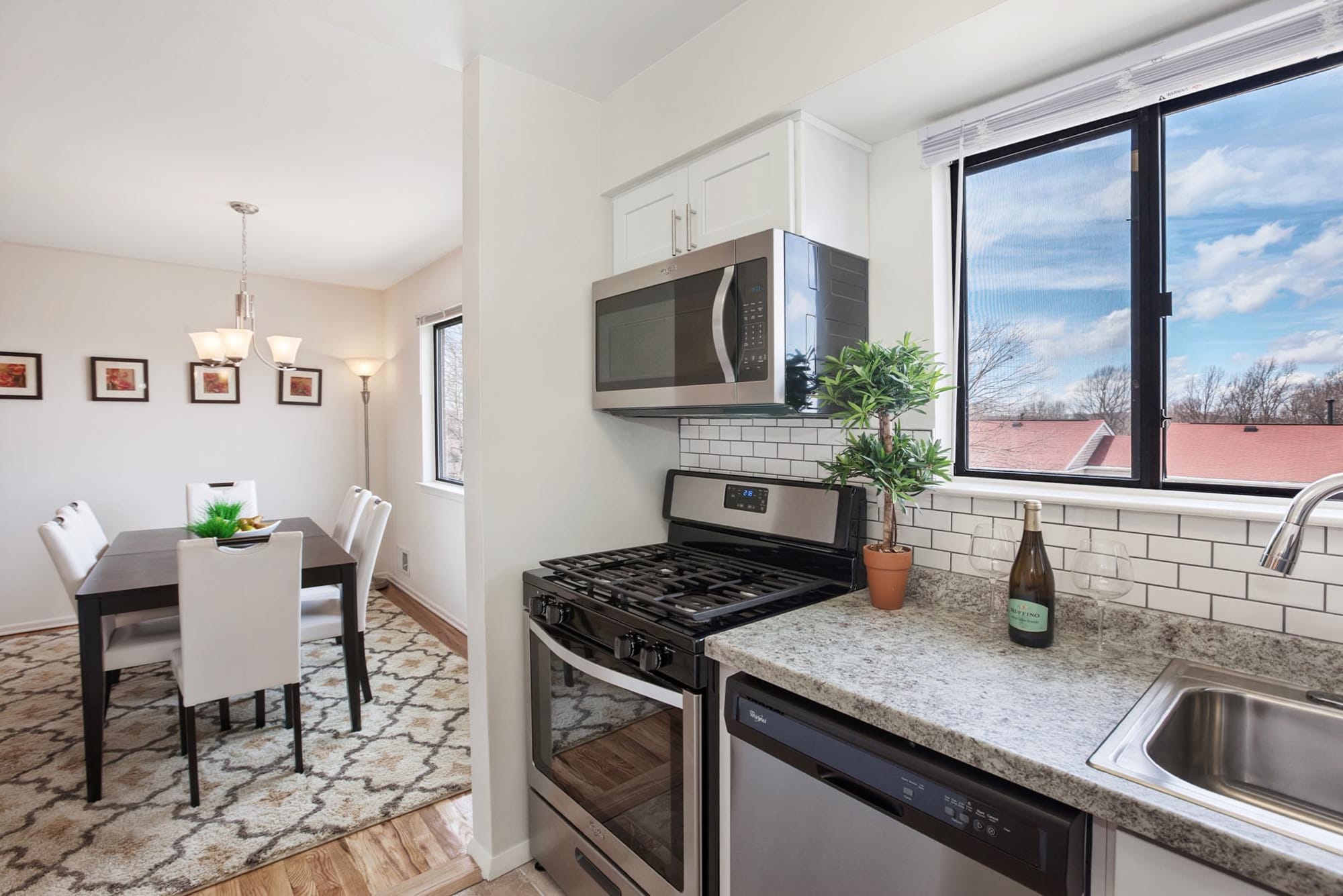 Staged kitchen with white subway tile, black grout, stainless steel appliances, sliding glass window, set in the background is a large dining room with hanging chandelier and another sliding glass window at Overlook at Flanders, Flanders, New Jersey