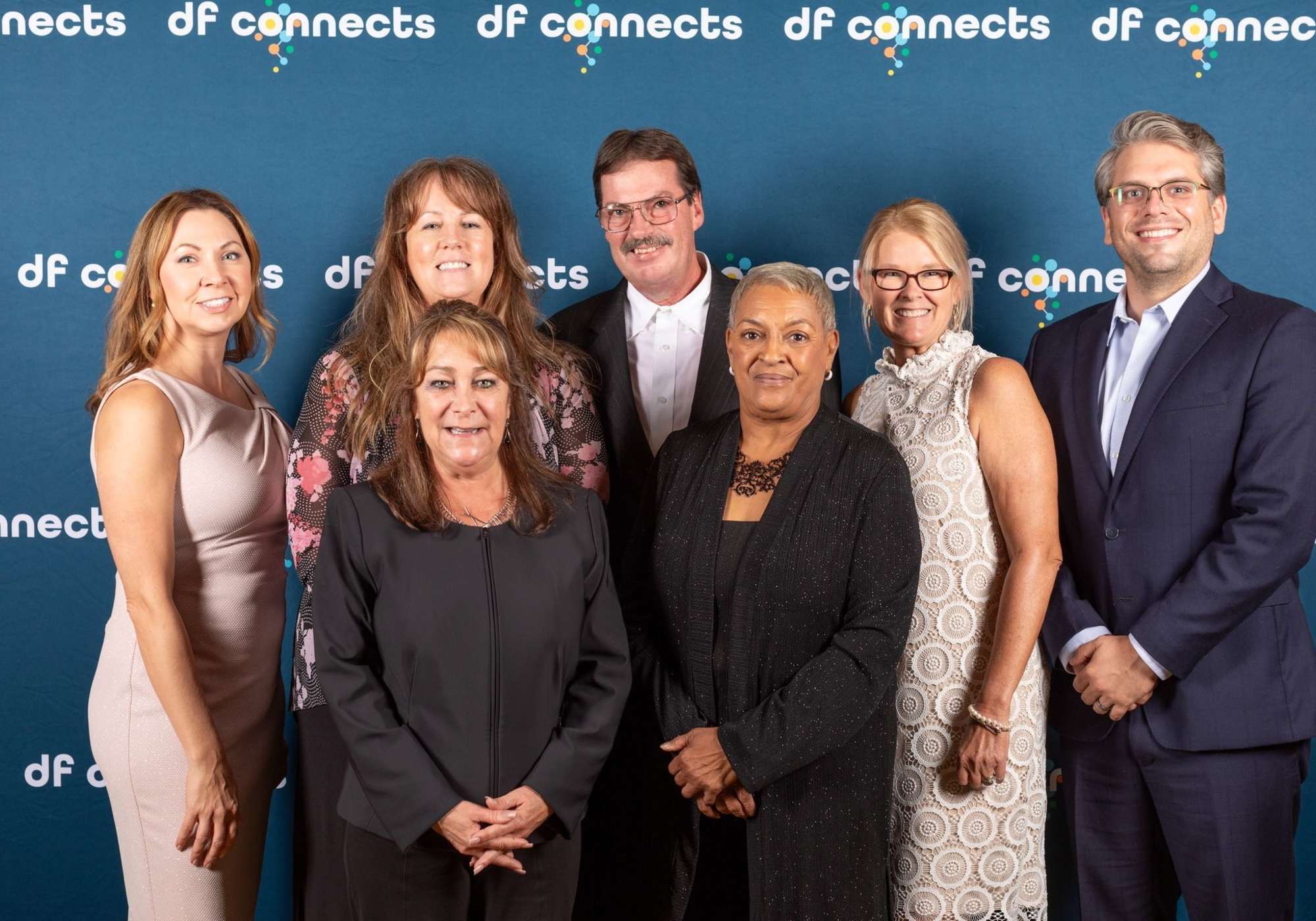A diverse group of people standing together in front of a vibrant blue wall, smiling and engaging with each other at Drucker + Falk in Newport News Virginia