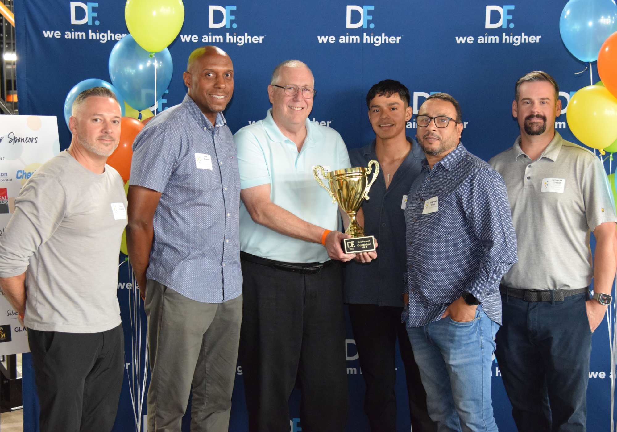 Five men proudly holding a trophy, surrounded by colorful balloons in a celebratory atmosphere at Drucker + Falk in Newport News Virginia