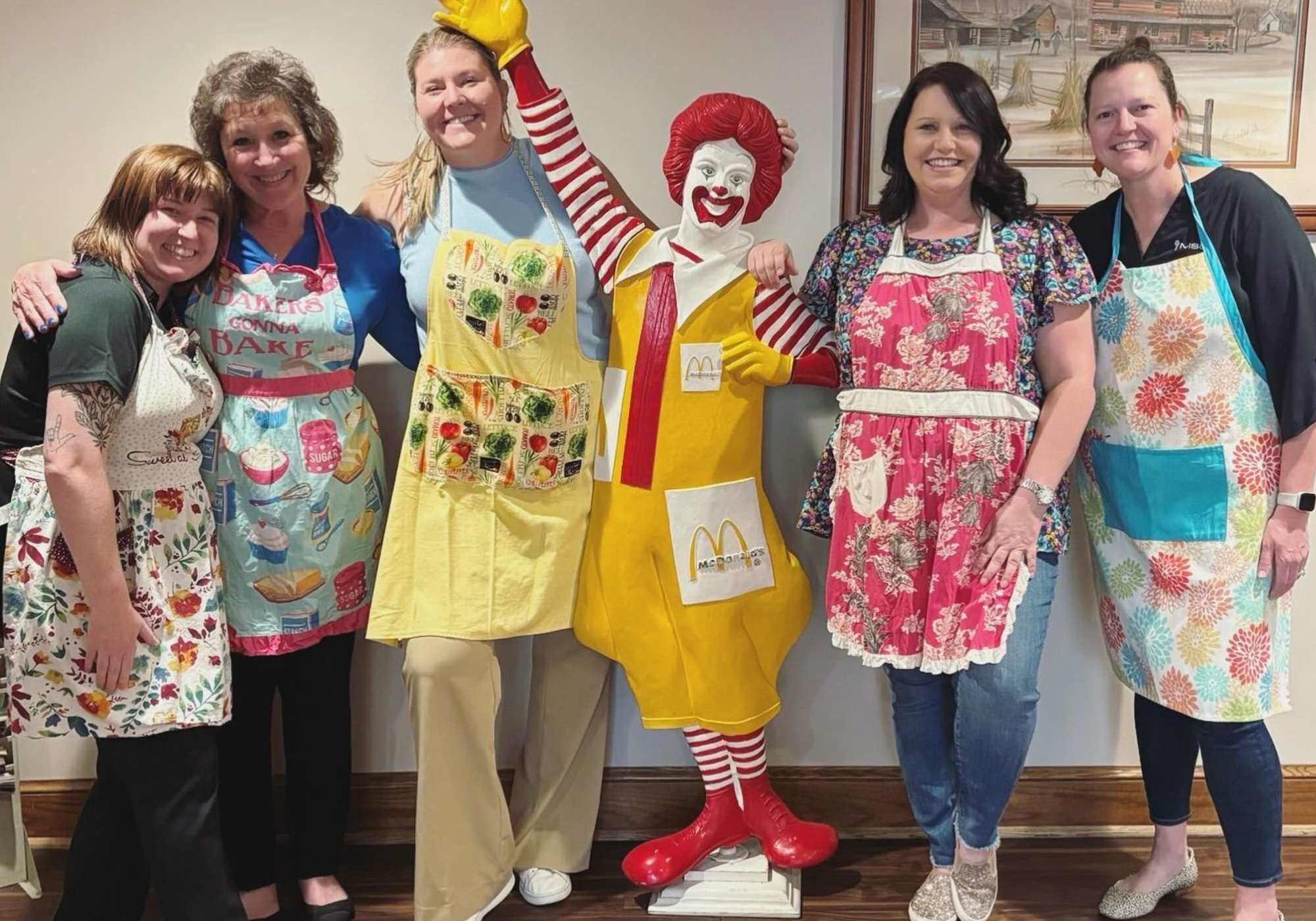 A group of women in aprons smiles while posing with a Ronald McDonald statue in a cheerful outdoor setting at Drucker + Falk in Newport News Virginia