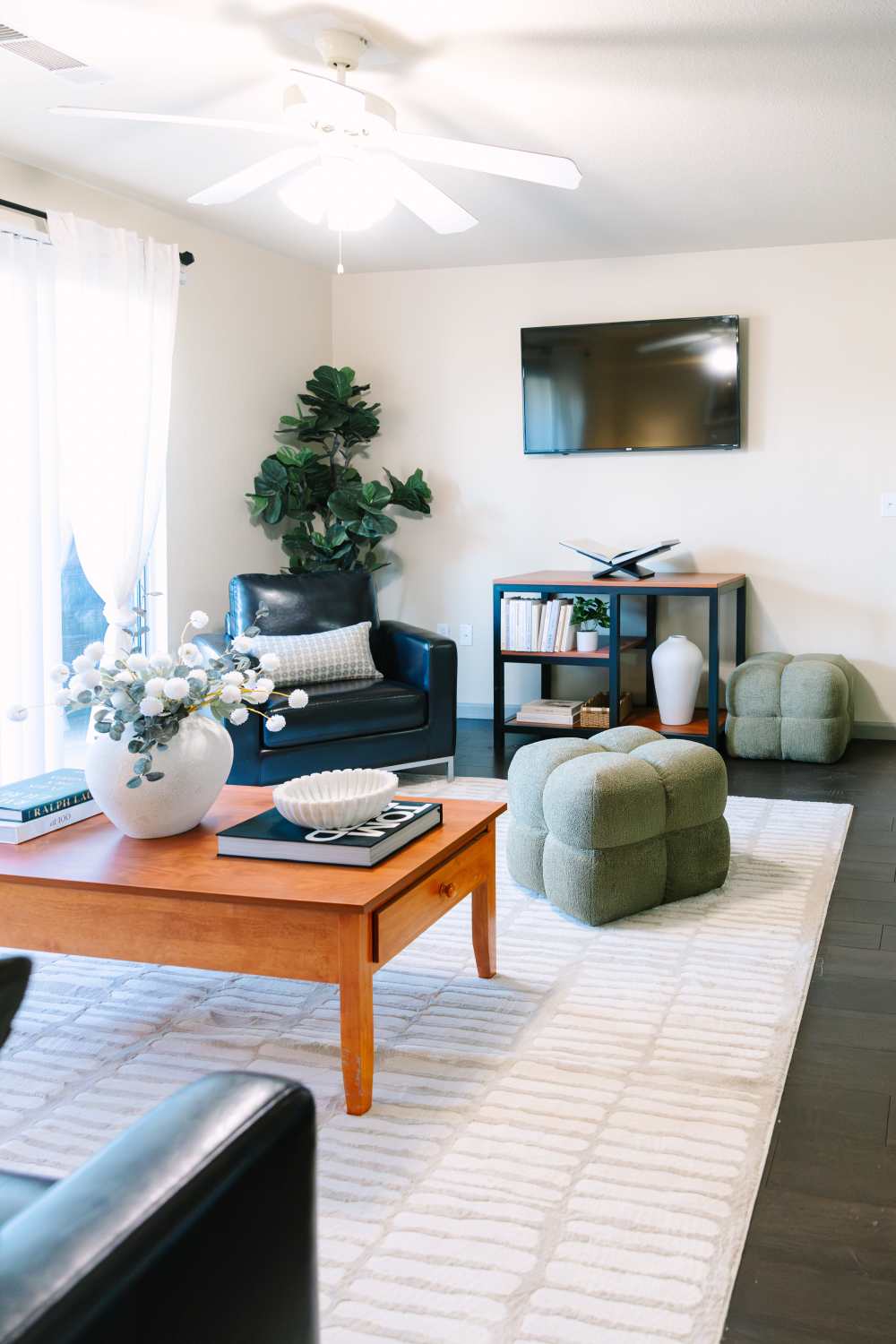  Modern living room with leather lounge chair, wall-mounted TV, wood coffee table, decorative ottomans, area rug, and large window with curtains at The Holly in Lubbock, Texas