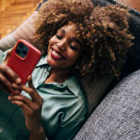 Woman relaxing on couch, smiling while using smartphone, cozy indoor setting with wood-style flooring at Retreat at the Park in Burlington, North Carolina