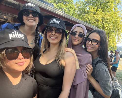 Group of friends wearing branded hats posing together outdoors near trees and a wooden structure at Pillar Properties in Seattle, Washington