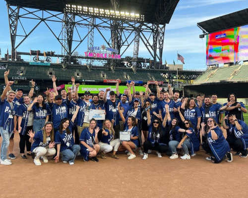 Large team in matching jerseys celebrating on a baseball field with stadium lights and signage behind them at Pillar Properties in Seattle, Washington
