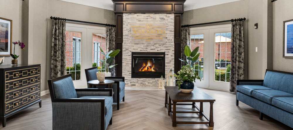 Cozy lounge with fireplace, armchairs, and large windows overlooking courtyard at Waltonwood Cherry Hill in Canton