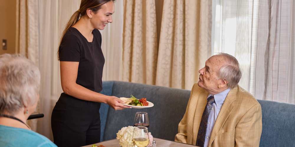 Server presenting a plate of food to an elderly man at a dining table at Barclay House of Oakleaf Village in Lexington, South Carolina