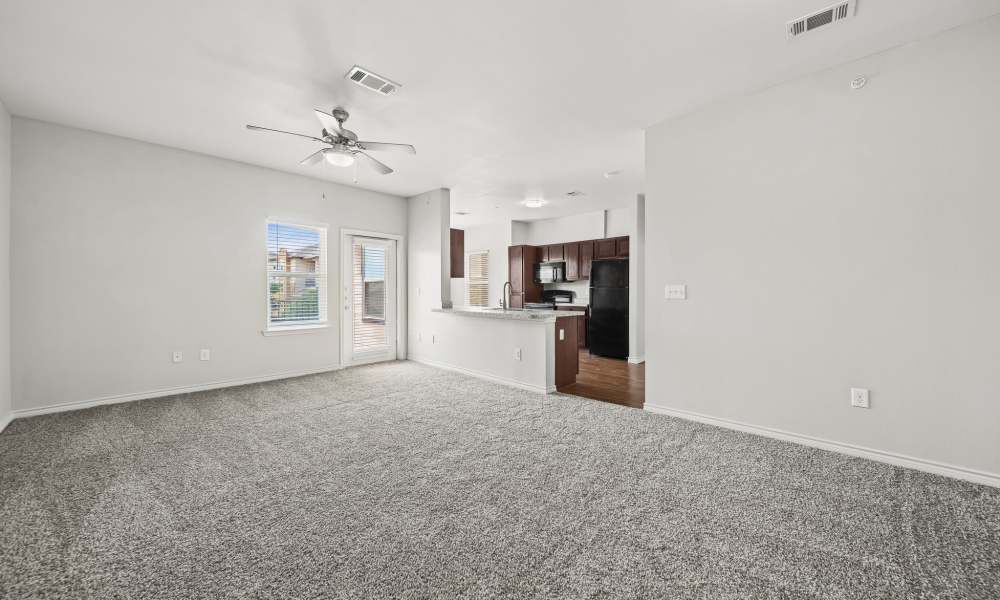 Unfurnished, carpeted living room with window, patio door, and kitchen in background at Sunset Lodge in Odessa, Texas