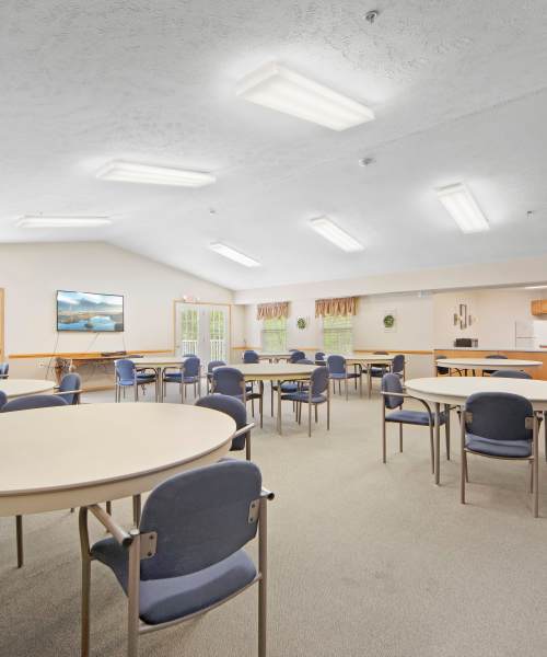 Spacious community room with round tables, chairs, and a kitchen area in the background at Heritage Village in Steubenville, Ohio