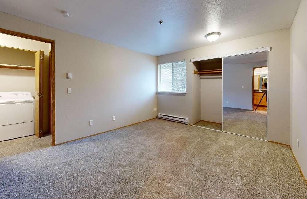 Spacious bedroom with a carpet, mirrored closet and a door leading to a laundry room with a washer and dryer at Heron Hills Apartment Homes in Gresham, Oregon