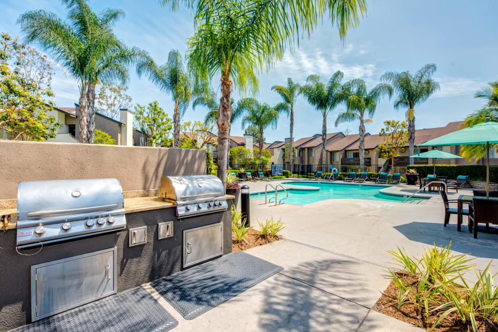 Barbecue area with comfortable seating and umbrellas aside the large swimming pool surrounded by lounge chairs and palm trees at Sofi Laguna Hills in Laguna Hills, California
