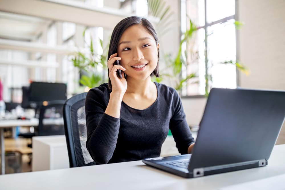 Young woman talking on the phone while sitting at a table with a laptop in front of her at a Jaybird Senior Living community