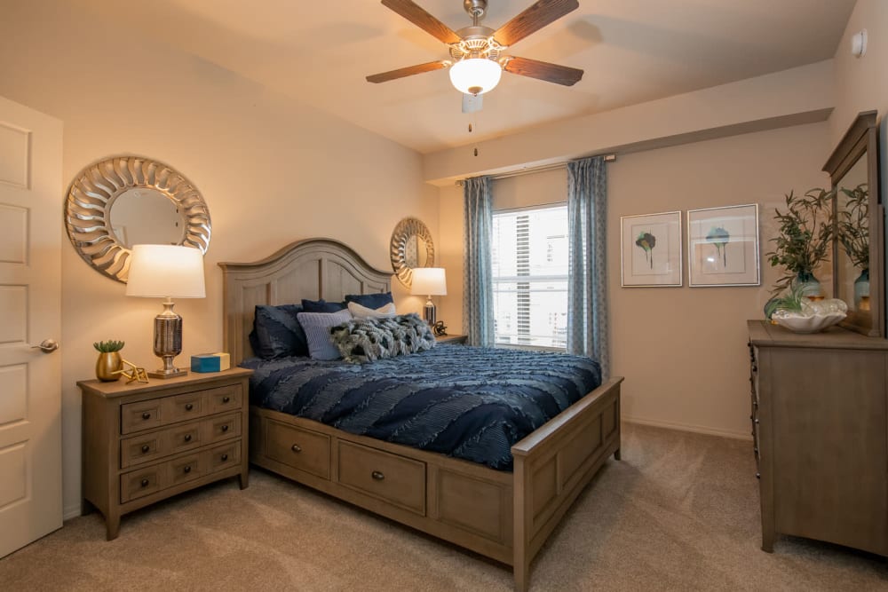 Plush carpeting, draped window, and a ceiling fan in the primary suite of a model apartment at Redbud Ranch Apartments in Broken Arrow, Oklahoma