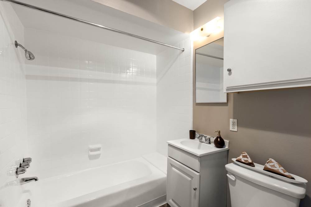  A bathroom interior with white bathtub with overhead shower, white toilet and stainless-steel faucet mounted on vanity cabinet at Reseda Village Green in Reseda, California