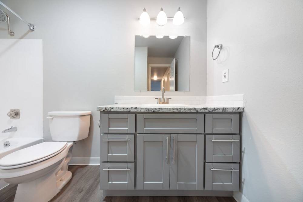 Bathroom with grey cabinetry, toilet, a mirror above the sink and a bathtub with a shower curtain rod at Lantera Apartments in Seattle, Washington