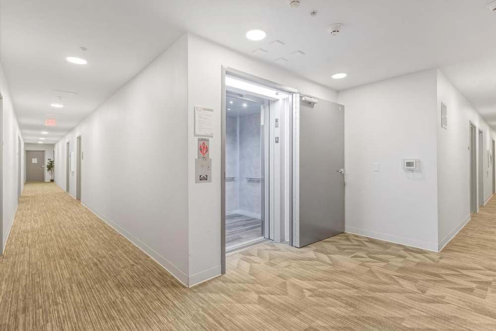 A modern elevator with gray doors opens into a well-lit hallway with beige carpeting at Koz on Killingsworth in Portland, Oregon