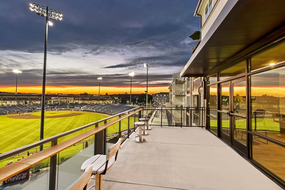A modern apartment balcony overlooking a professional baseball stadium at sunset, with stadium lights glowing, a vivid orange and blue sky, and sleek patio seating along the railing.