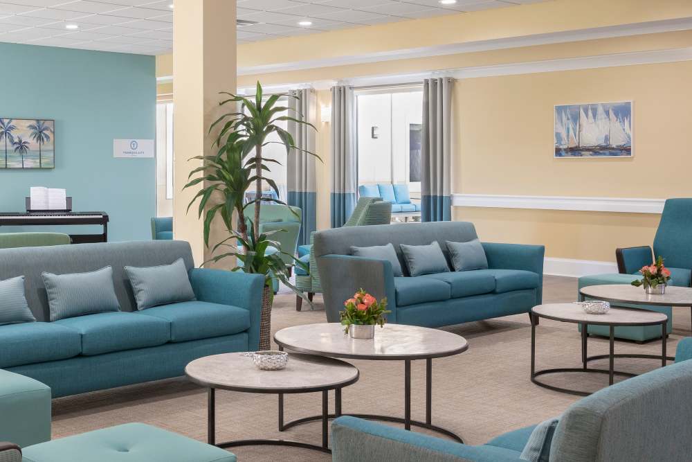 Lounge with teal sofas, marble-top tables, flower arrangements, and sailboat-themed wall art at The Pineapple House at Sapphire Lakes in Naples, Florida
