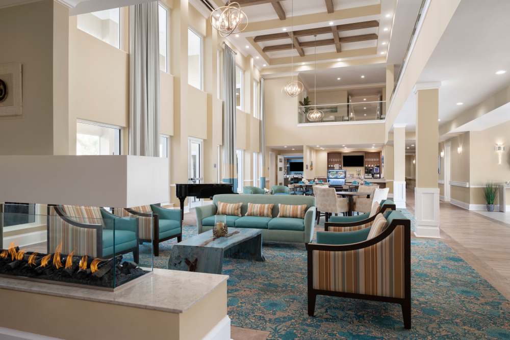 Elegant living area with blue and beige seating, fireplace, grand piano, and high ceilings with wooden beams at The Pineapple House at Sapphire Lakes in Naples, Florida