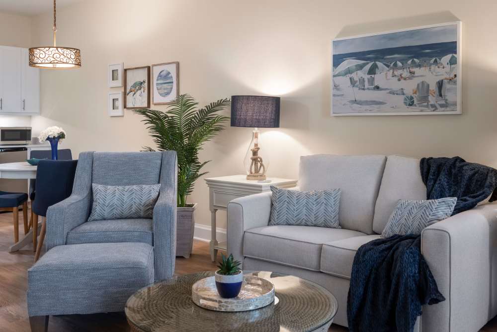 Cozy living room with sofa, armchair, glass coffee table, snowy landscape art, and a kitchenette in the background at The Pineapple House at Sapphire Lakes in Naples, Florida