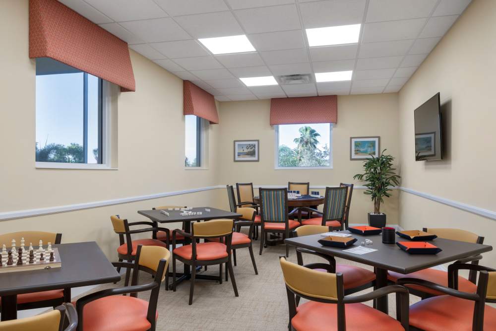 Activity room with tables set for games, red valances on windows, wall art, and a mounted TV at The Pineapple House at Sapphire Lakes in Naples, Florida