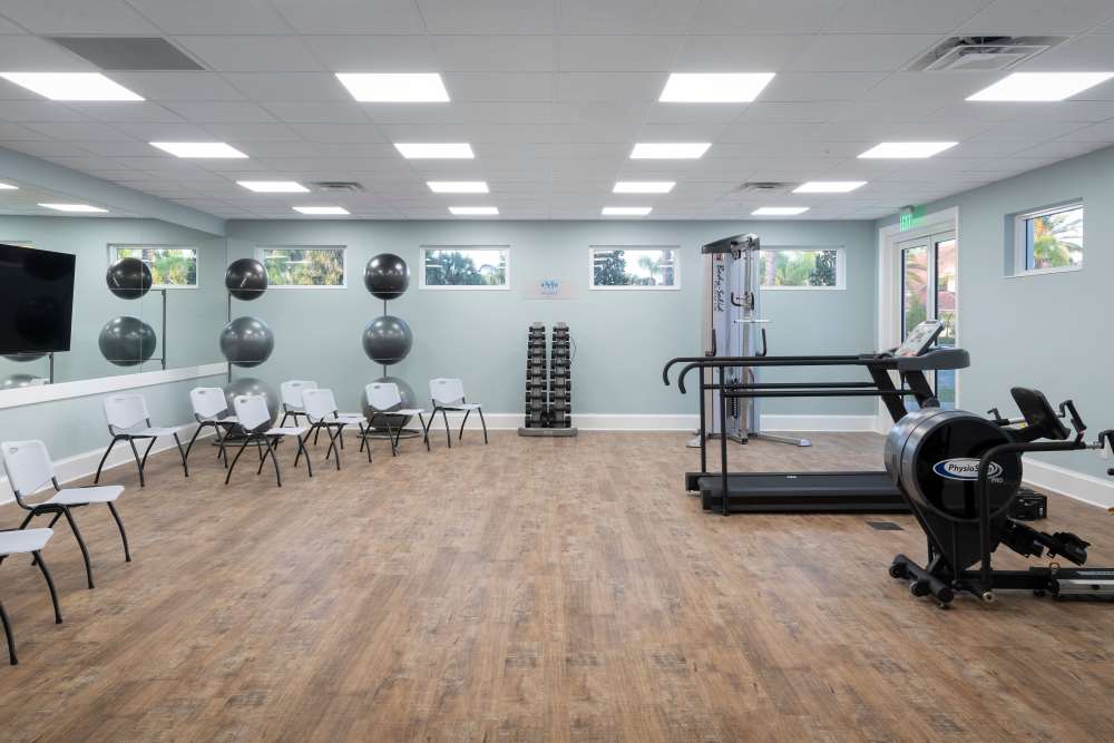 Fitness room with treadmills, bikes, dumbbells, chairs, wall mirror, and natural light from windows at The Pineapple House at Sapphire Lakes in Naples, Florida