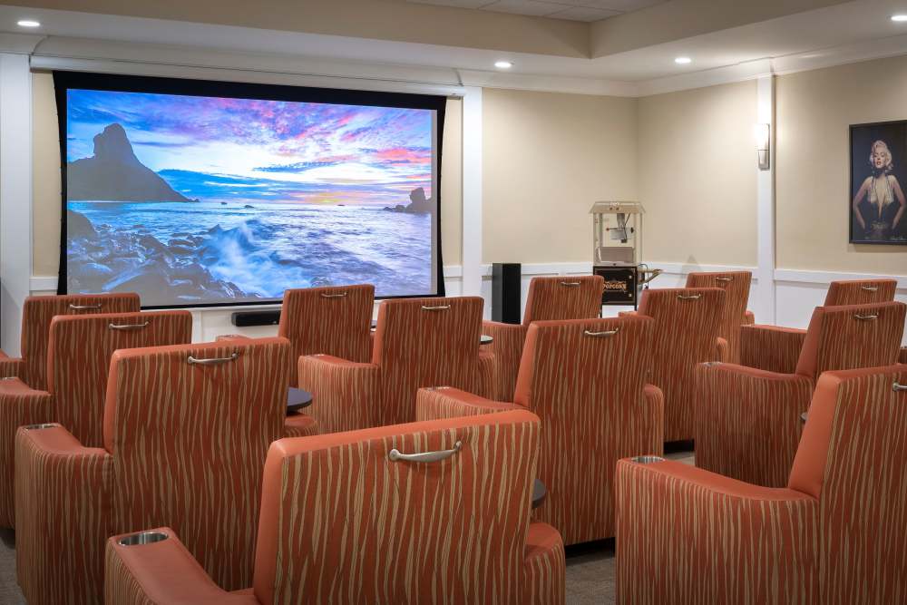 Small theater room with orange chairs, large screen showing ocean scene, and a popcorn machine in the back at The Pineapple House at Sapphire Lakes in Naples, Florida