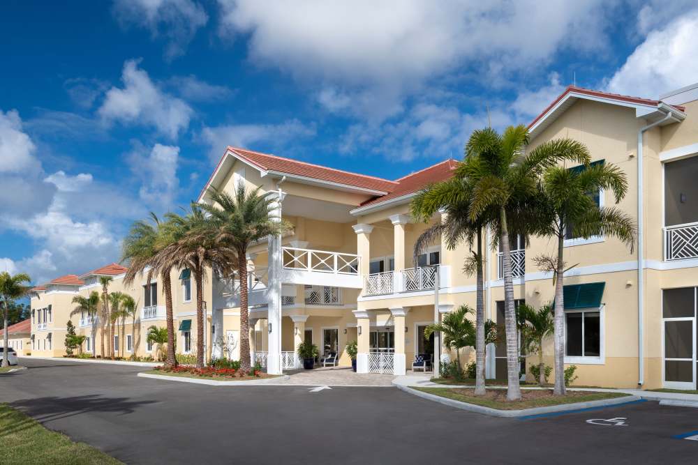 Two-story yellow building with red-tiled roof, balconies, palm trees, and a paved driveway at The Pineapple House at Sapphire Lakes in Naples, Florida