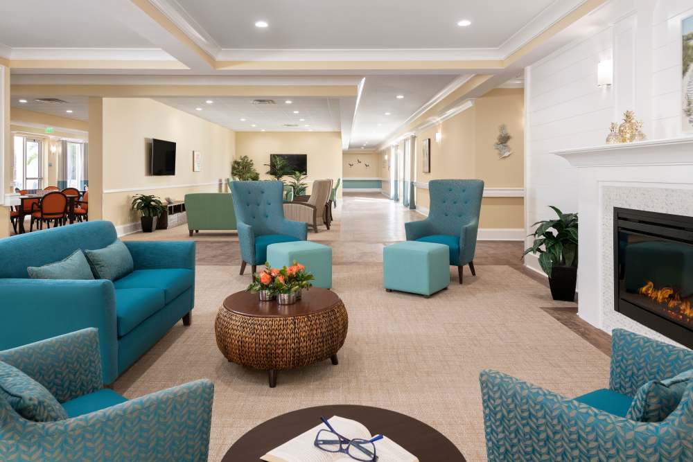 Modern lounge with blue chairs, wicker coffee table, fireplace, large windows, and beige walls at The Pineapple House at Sapphire Lakes in Naples, Florida