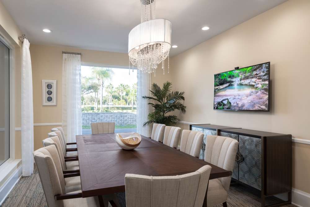 Modern dining room with wooden table, chandelier, upholstered chairs, large window, and flat-screen TV at The Pineapple House at Sapphire Lakes in Naples, Florida