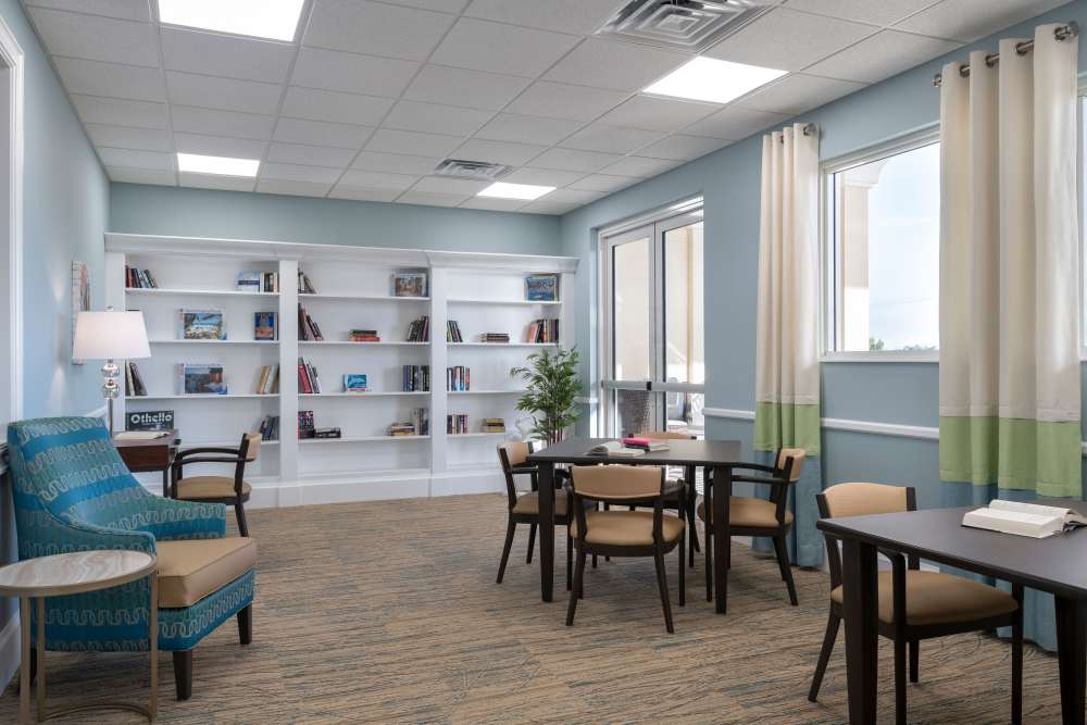 Modern reading room with bookshelves, tables, chairs, large windows, and a cozy armchair in the corner at The Pineapple House at Sapphire Lakes in Naples, Florida